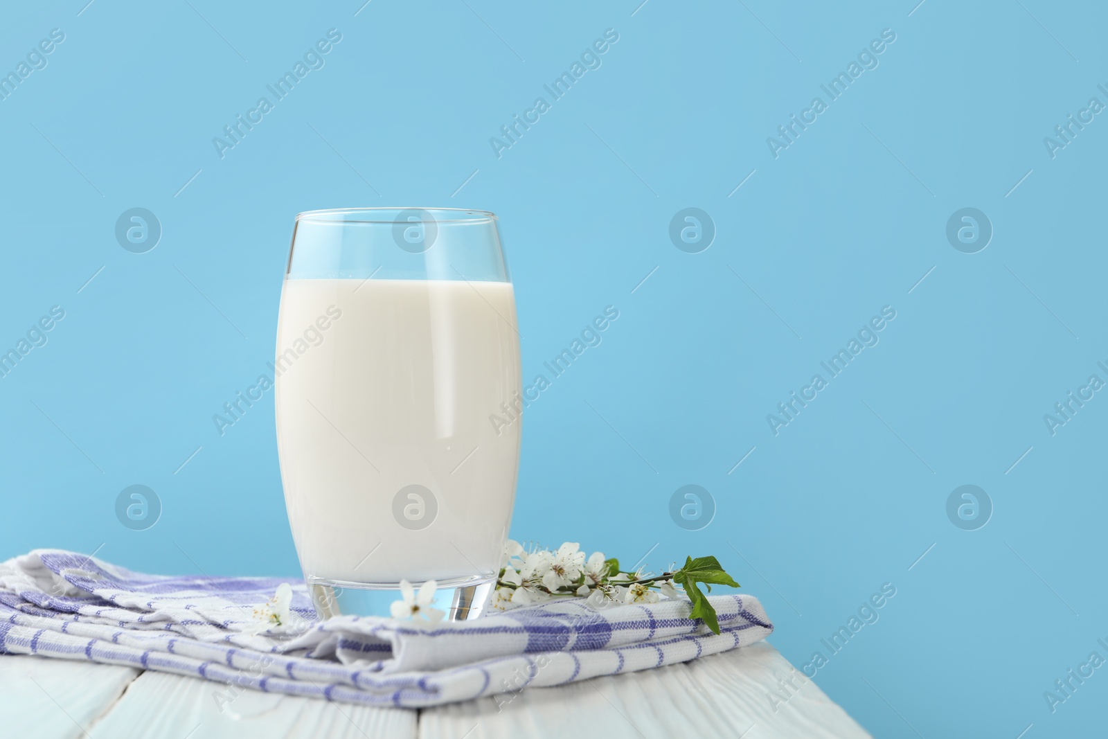 Photo of Fresh milk in glass and blossoms on white wooden table against light blue background, closeup