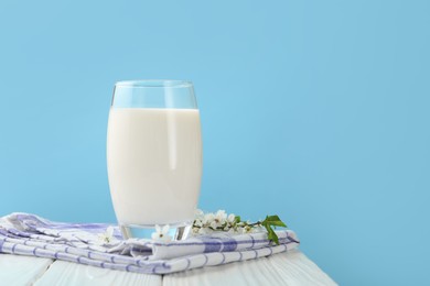 Photo of Fresh milk in glass and blossoms on white wooden table against light blue background, closeup