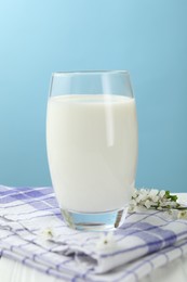 Photo of Fresh milk in glass and blossoms on white wooden table against light blue background, closeup