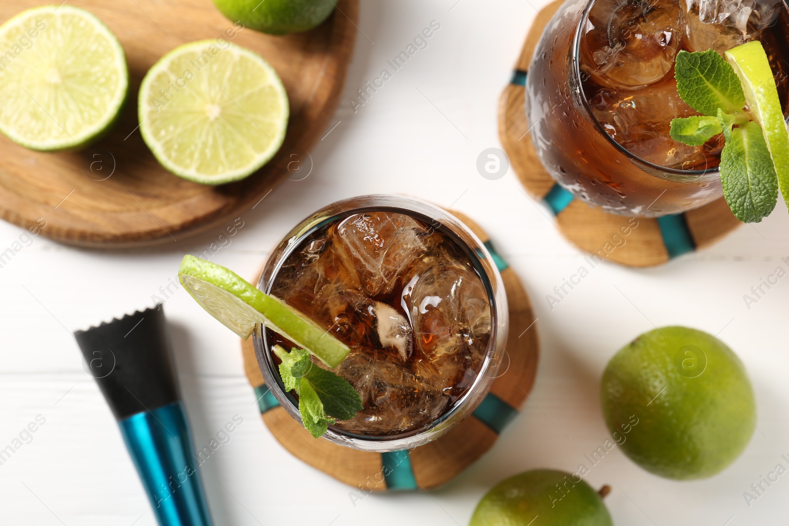 Tasty Cuba Libre cocktail in glasses, limes and muddler on white table, flat lay Photo of Tasty Cuba Libre cocktail in glasses, limes and muddler on white table, flat lay
