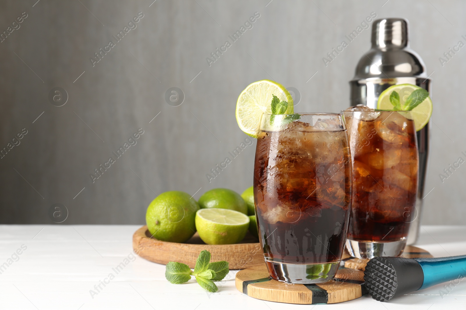 Tasty Cuba Libre cocktail in glasses, ingredients, muddler and shaker on white wooden table, space for text Photo of Tasty Cuba Libre cocktail in glasses, ingredients, muddler and shaker on white wooden table, space for text