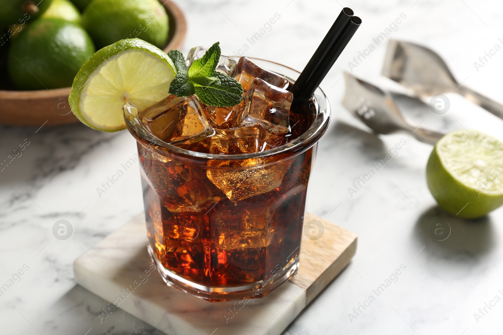 Tasty Cuba Libre cocktail in glass, limes and tongs on white marble table, closeup Photo of Tasty Cuba Libre cocktail in glass, limes and tongs on white marble table, closeup