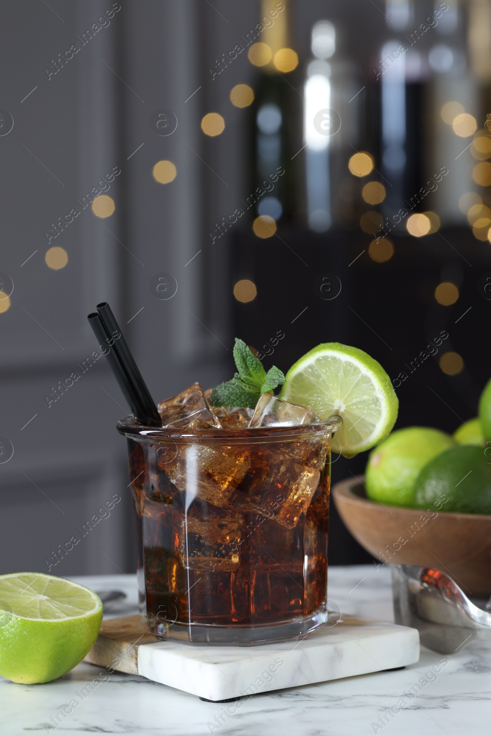 Tasty Cuba Libre cocktail in glass, limes and tongs on white marble table, bokeh effect Photo of Tasty Cuba Libre cocktail in glass, limes and tongs on white marble table, bokeh effect