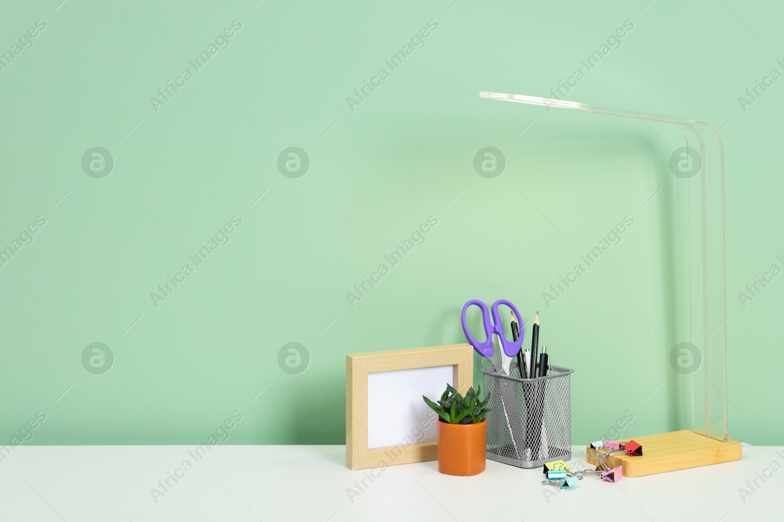 Stylish workplace with lamp, stationery and plant on white desk near light green wall. Space for text Photo of Stylish workplace with lamp, stationery and plant on white desk near light green wall. Space for text