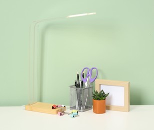 Photo of Stylish workplace with lamp, stationery and plant on white desk near light green wall