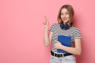 Photo of Portrait of smiling teenage girl with book pointing at something on pink background. Space for text