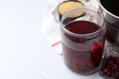 Photo of Tasty cherry compot in glass jar and ingredients on white tiled table, closeup. Space for text