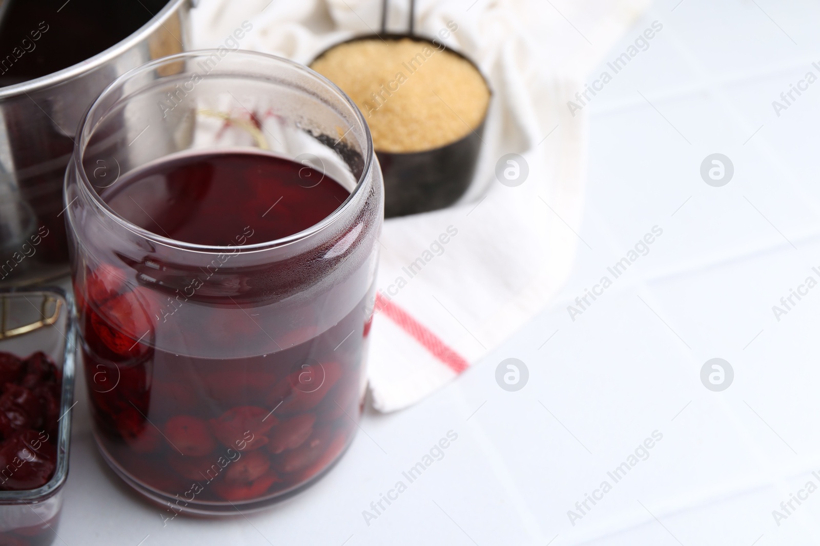 Photo of Tasty cherry compot in glass jar and ingredients on white tiled table, closeup. Space for text