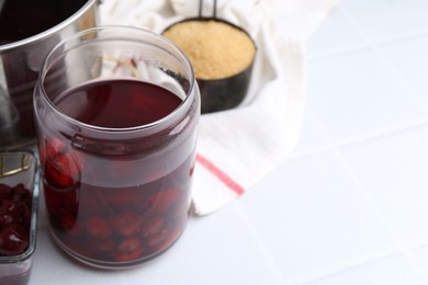 Photo of Tasty cherry compot in glass jar and ingredients on white tiled table, closeup. Space for text