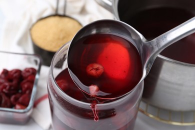 Photo of Pouring tasty cherry compot into glass jar on white table, closeup