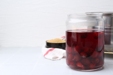 Photo of Tasty cherry compot in glass jar on white tiled table, closeup. Space for text