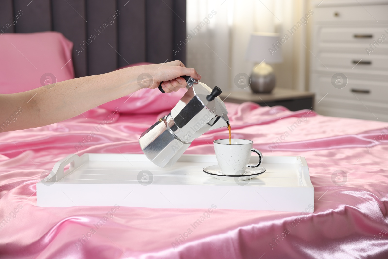 Woman pouring coffee from moka pot into cup on bed with silk linens, closeup Photo of Woman pouring coffee from moka pot into cup on bed with silk linens, closeup