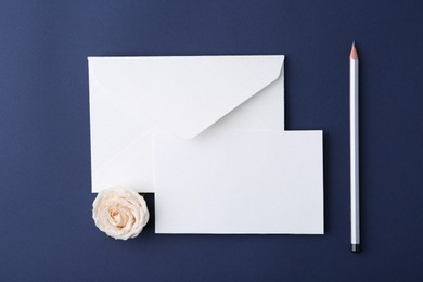 Photo of Blank greeting card, paper envelope, rose and pencil on dark blue background, top view