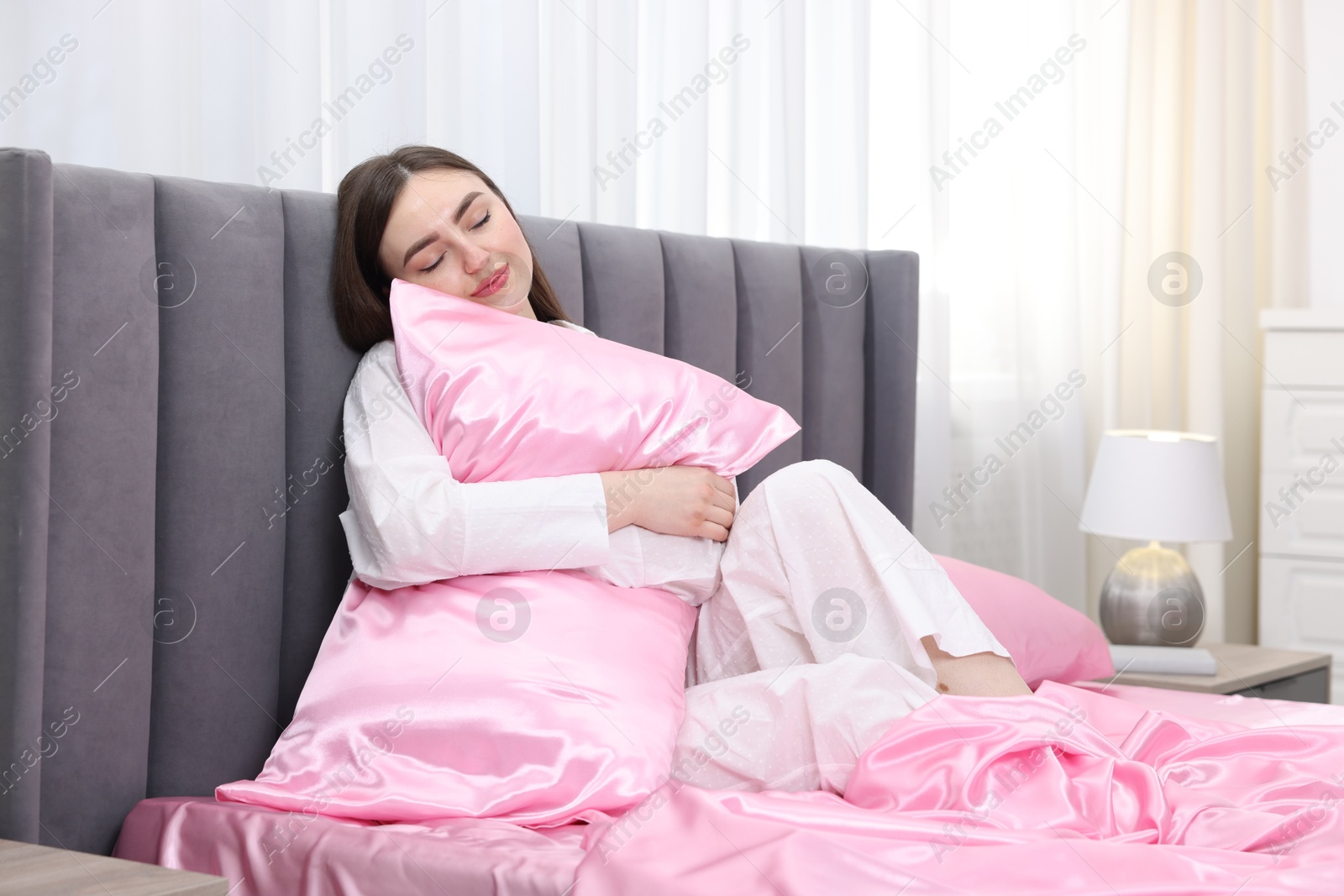 Woman hugging pillow on bed with pink silk linens indoors Photo of Woman hugging pillow on bed with pink silk linens indoors
