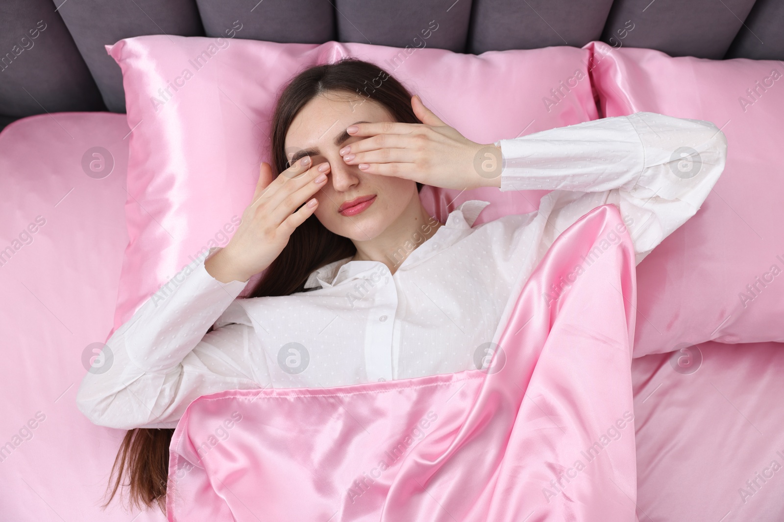 Woman covering eyes with hands in bed with pink silk linens, top view Photo of Woman covering eyes with hands in bed with pink silk linens, top view
