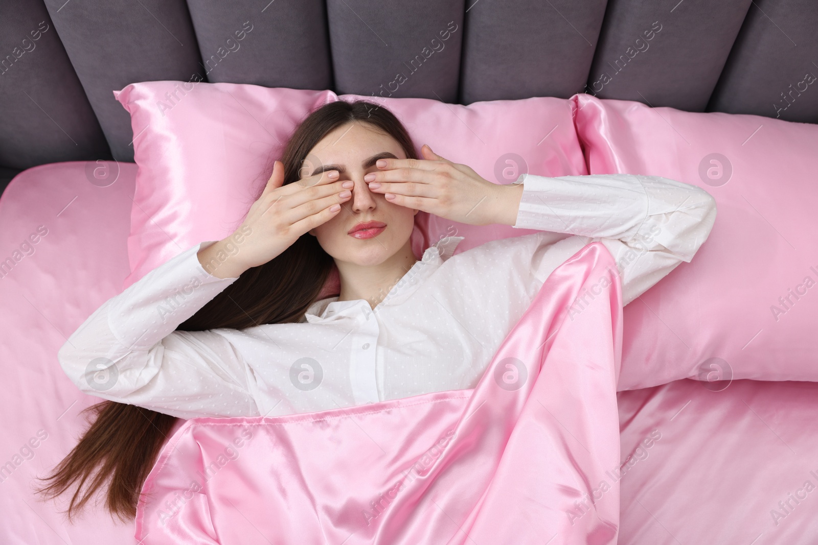 Woman covering eyes with hands in bed with pink silk linens, top view Photo of Woman covering eyes with hands in bed with pink silk linens, top view