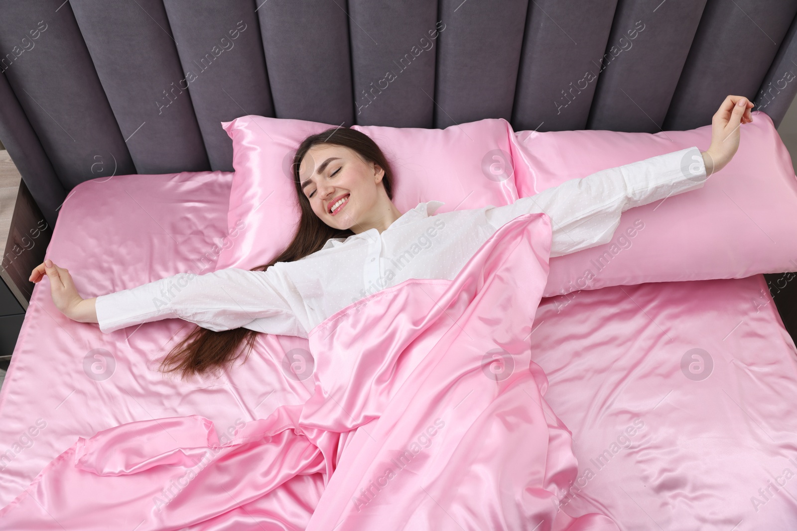 Smiling woman stretching in bed with pink silk linens indoors, above view Photo of Smiling woman stretching in bed with pink silk linens indoors, above view