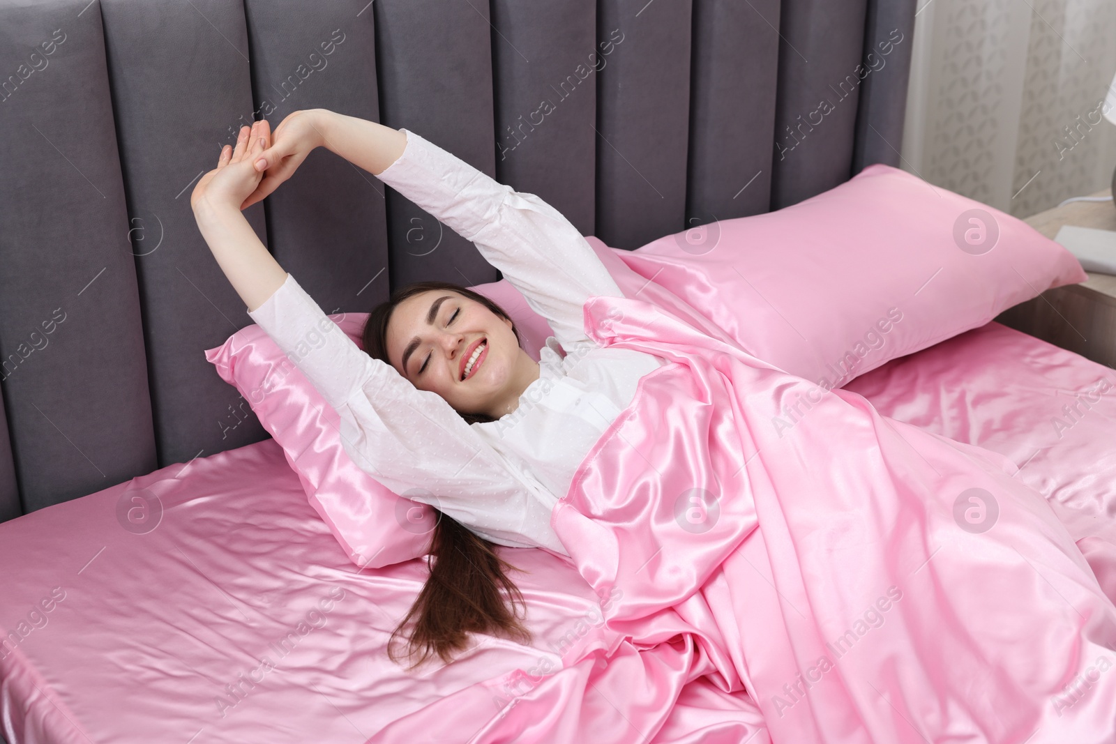 Smiling woman stretching in bed with pink silk linens indoors Photo of Smiling woman stretching in bed with pink silk linens indoors