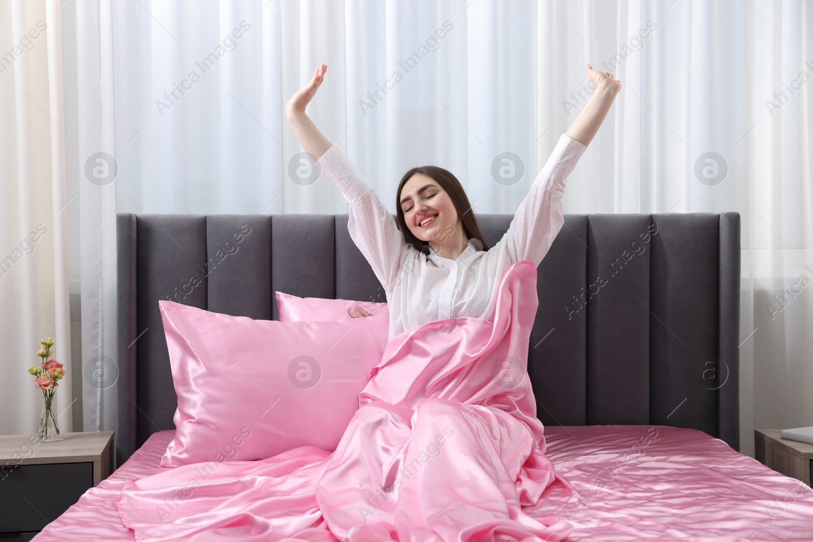 Smiling woman stretching in bed with pink silk linens indoors Photo of Smiling woman stretching in bed with pink silk linens indoors