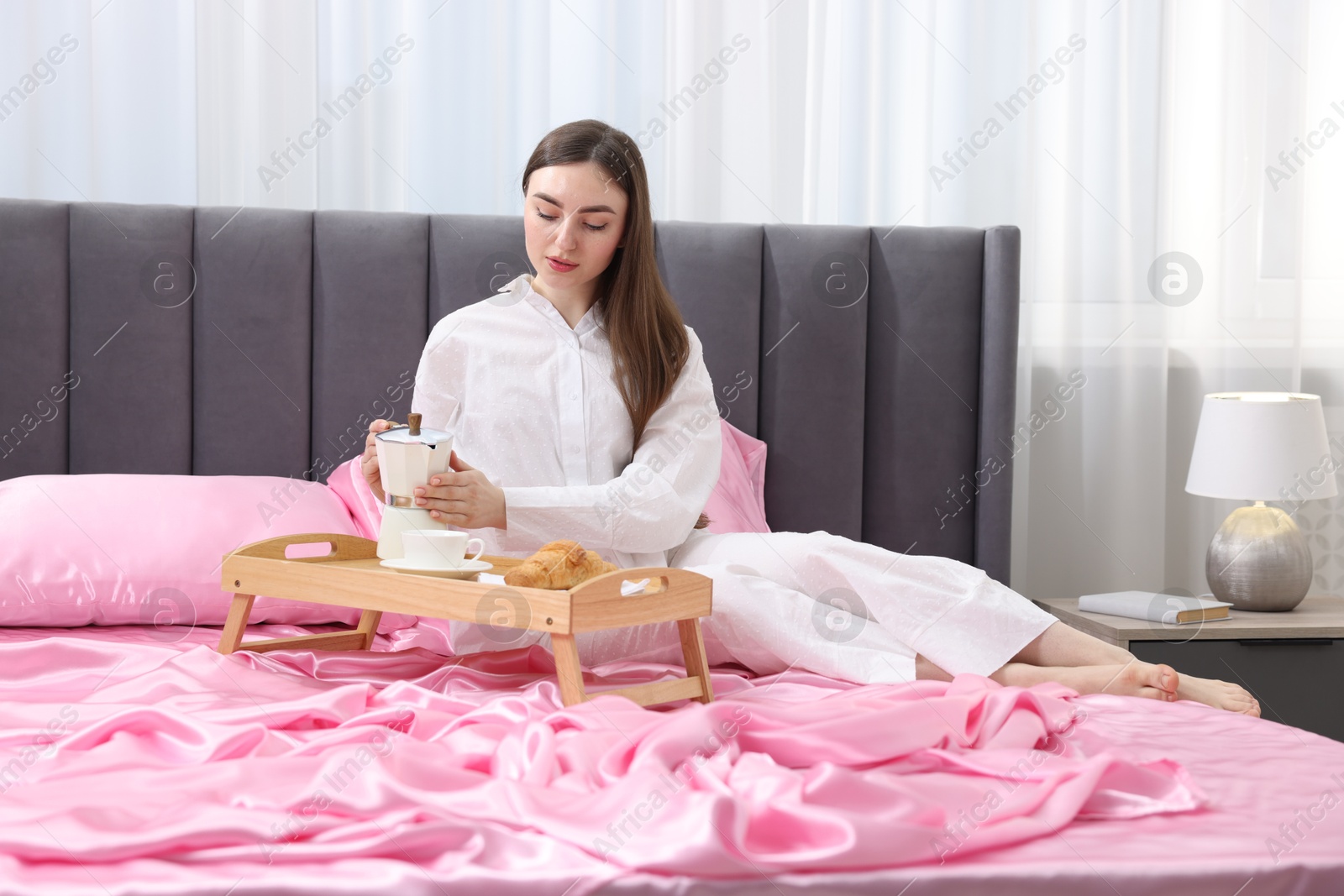 Woman pouring coffee from moka pot into cup on bed with pink silk linens indoors Photo of Woman pouring coffee from moka pot into cup on bed with pink silk linens indoors