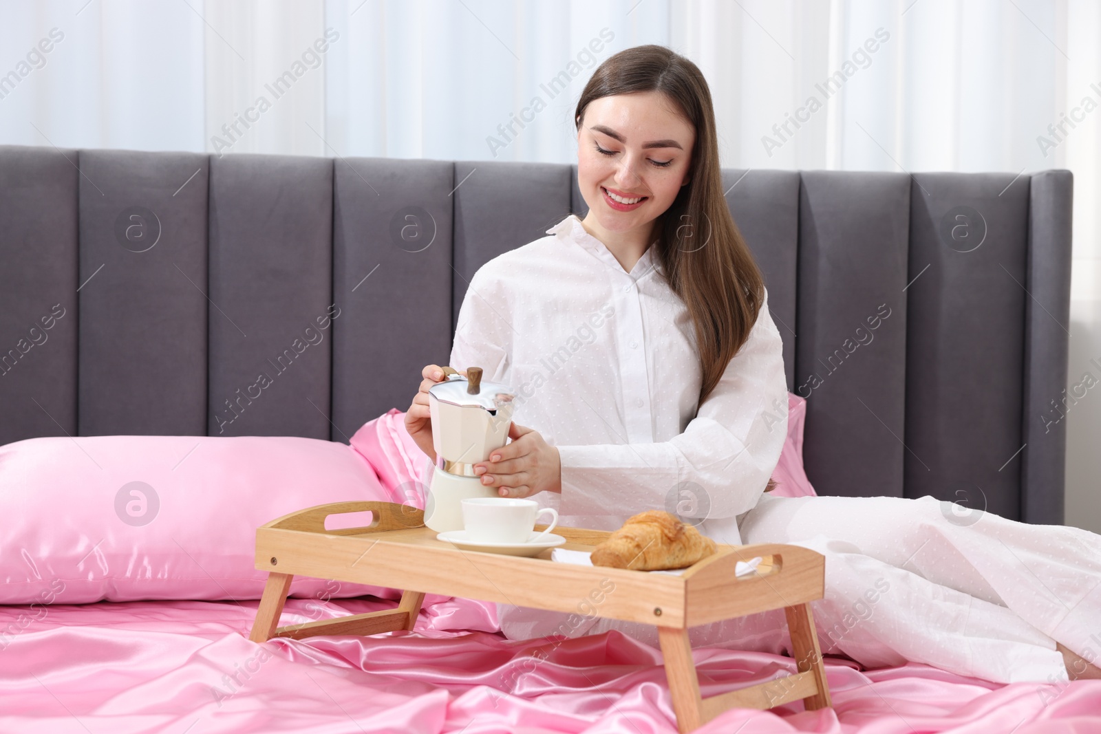 Smiling woman pouring coffee from moka pot into cup on bed with pink silk linens indoors Photo of Smiling woman pouring coffee from moka pot into cup on bed with pink silk linens indoors
