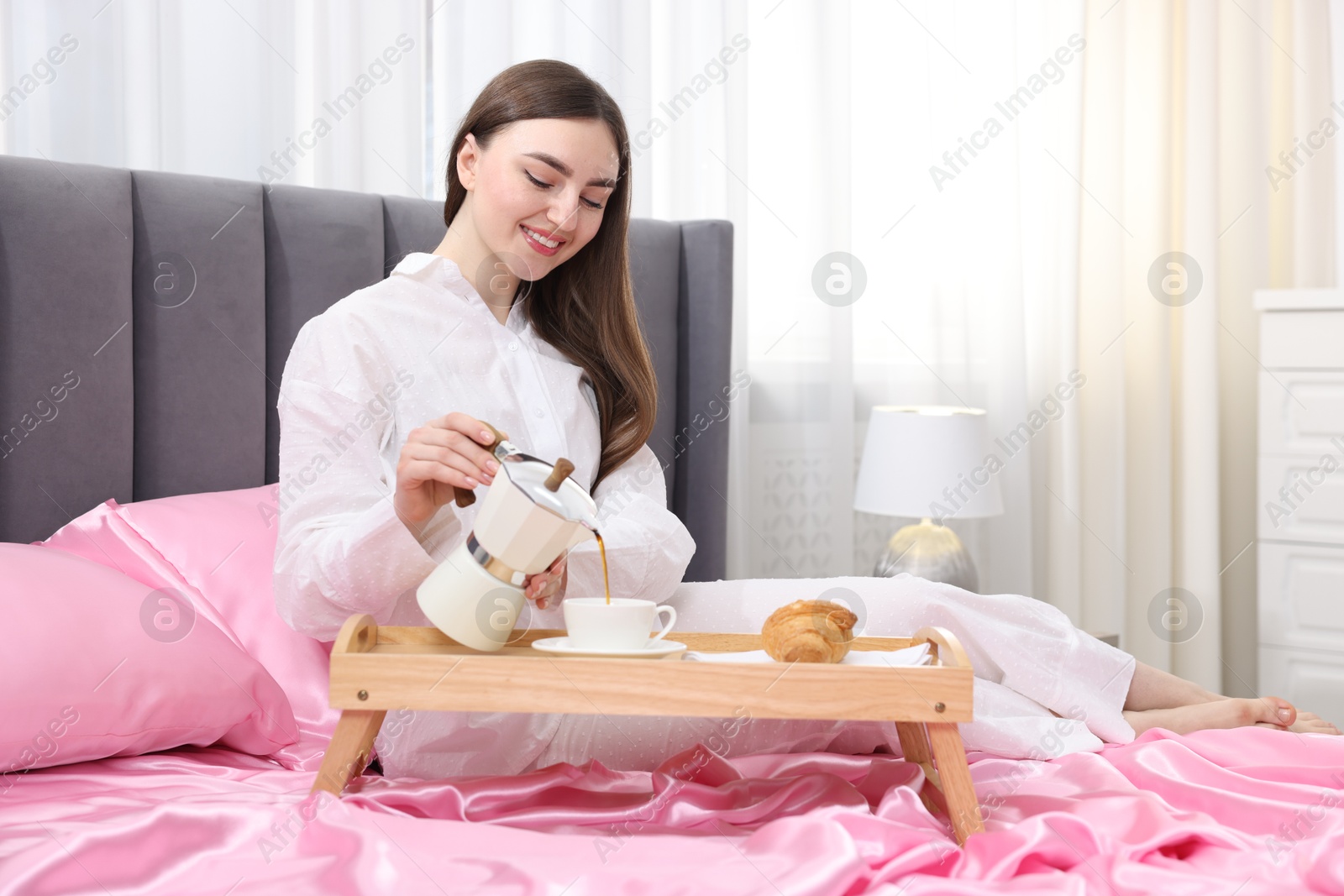 Smiling woman pouring coffee from moka pot into cup on bed with pink silk linens indoors Photo of Smiling woman pouring coffee from moka pot into cup on bed with pink silk linens indoors