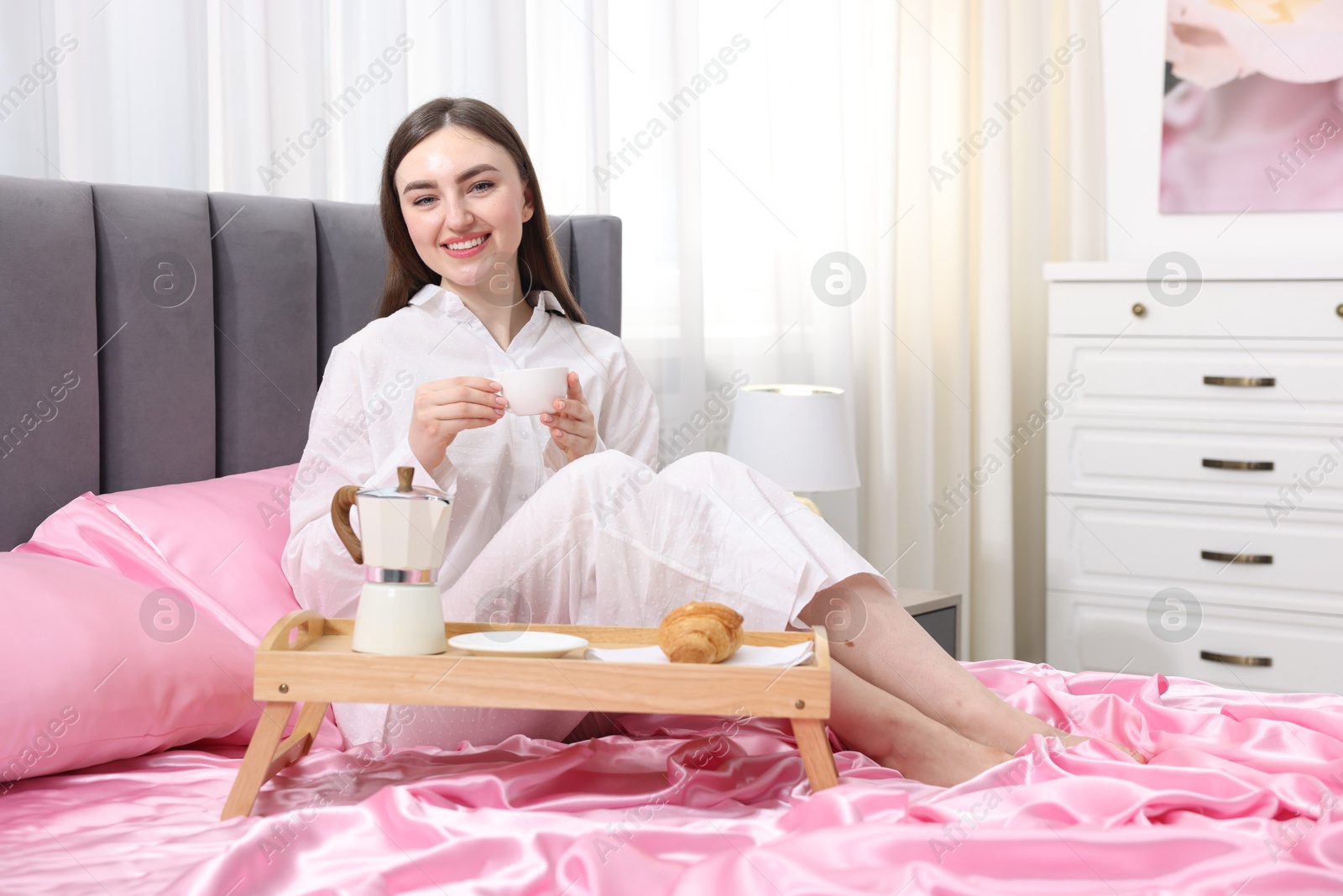 Smiling woman with cup of coffee on bed with pink silk linens indoors Photo of Smiling woman with cup of coffee on bed with pink silk linens indoors