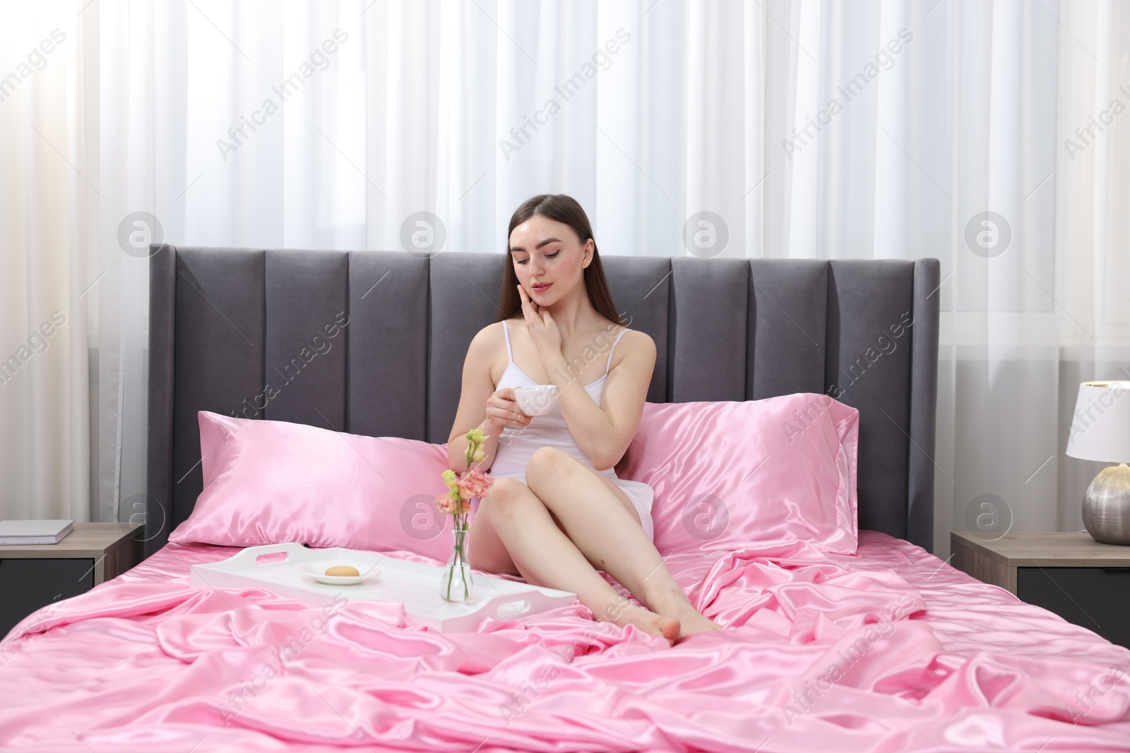 Woman with cup of coffee on bed with pink silk linens indoors Photo of Woman with cup of coffee on bed with pink silk linens indoors
