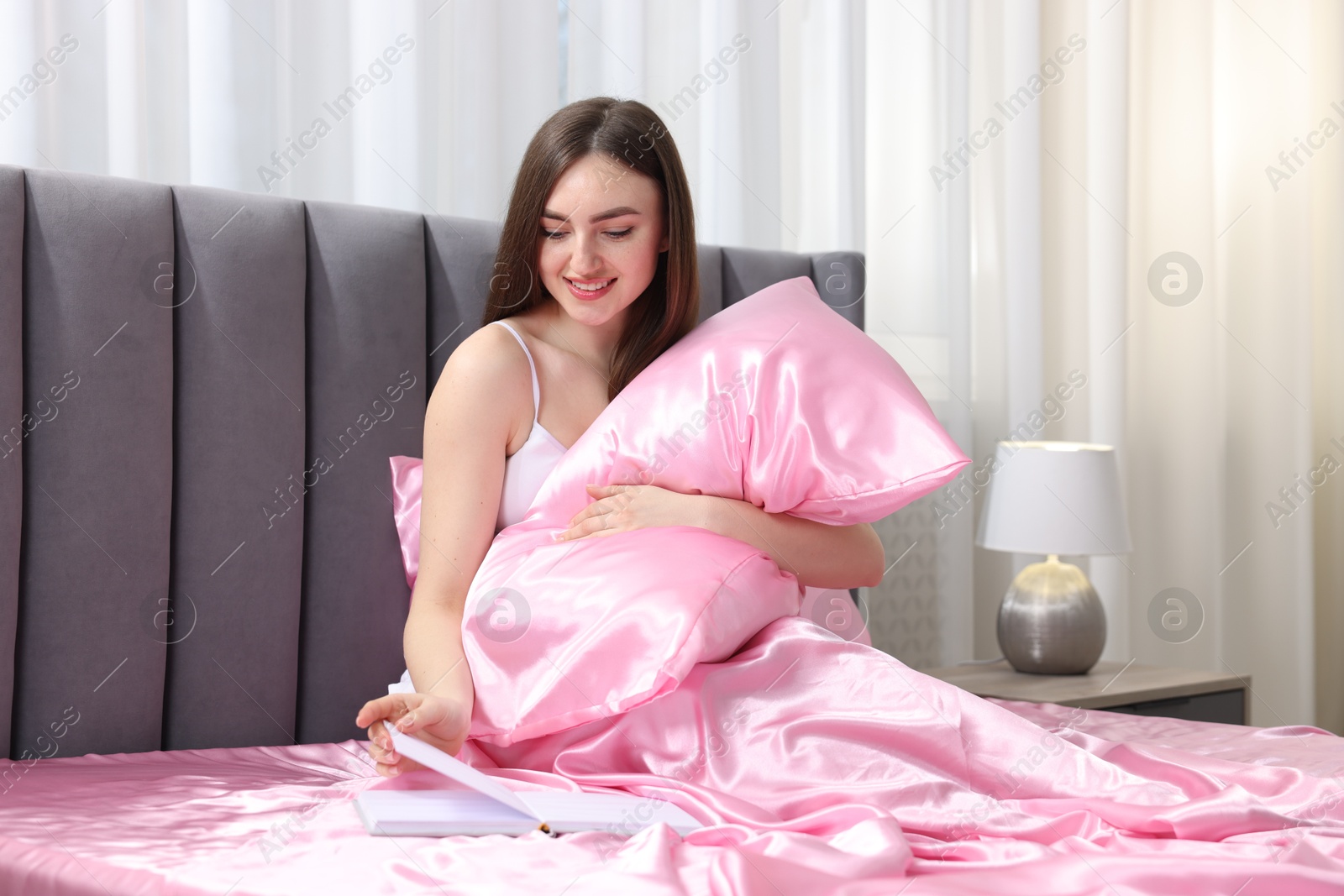 Smiling woman reading book in bed with pink silk linens indoors Photo of Smiling woman reading book in bed with pink silk linens indoors