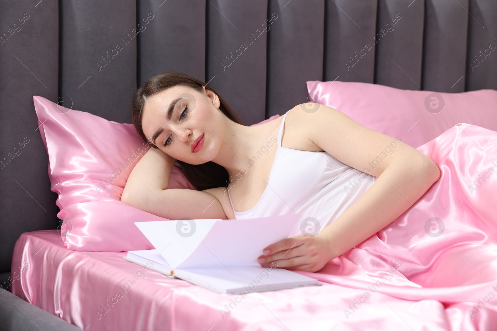 Woman reading book in bed with pink silk linens indoors Photo of Woman reading book in bed with pink silk linens indoors