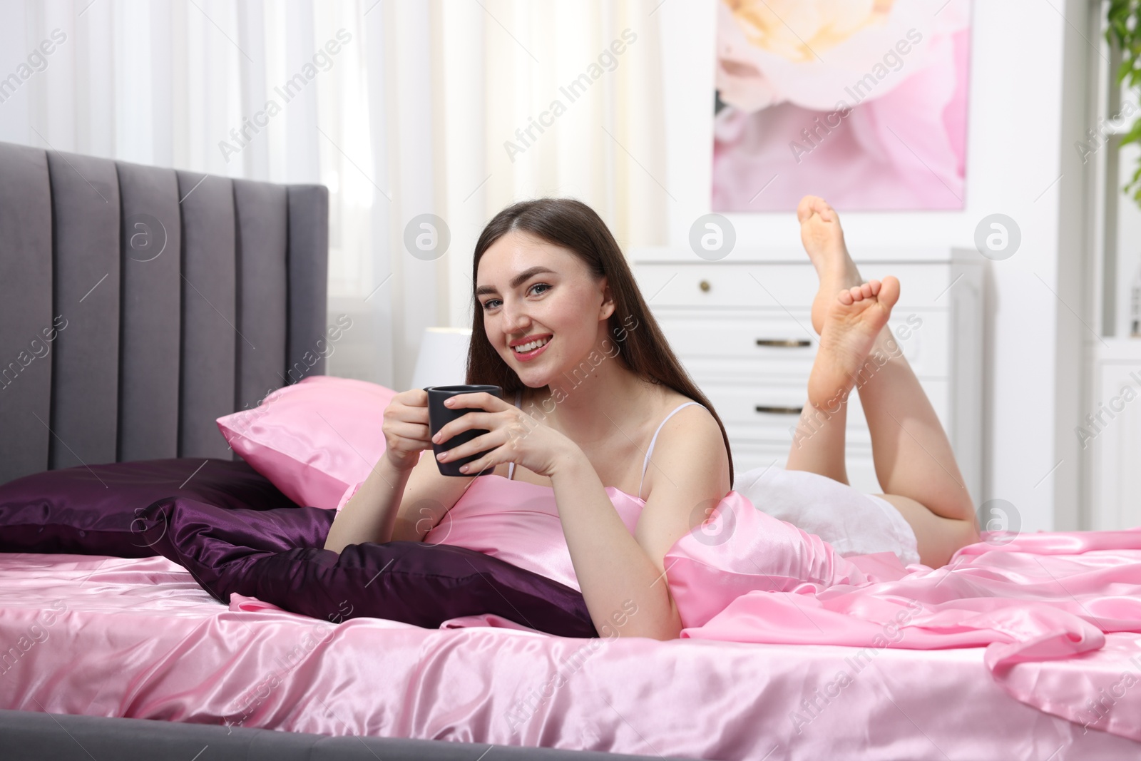 Smiling woman with cup of hot drink on bed with pink silk linens indoors Photo of Smiling woman with cup of hot drink on bed with pink silk linens indoors