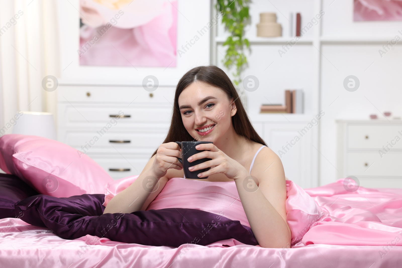 Smiling woman with cup of hot drink on bed with pink silk linens indoors Photo of Smiling woman with cup of hot drink on bed with pink silk linens indoors