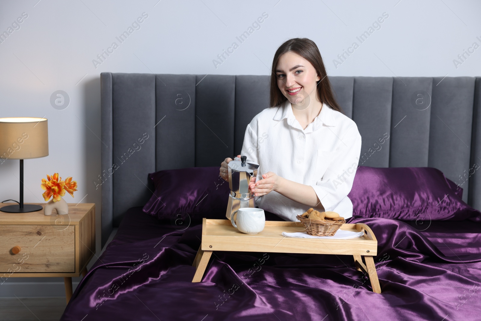 Smiling woman pouring coffee from moka pot into cup on bed with purple silk linens indoors Photo of Smiling woman pouring coffee from moka pot into cup on bed with purple silk linens indoors