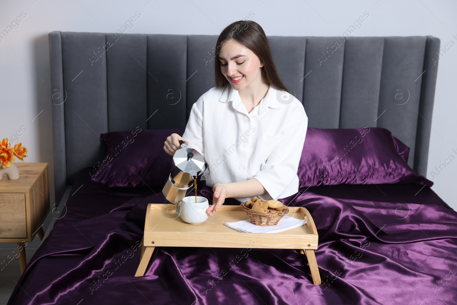 Smiling woman pouring coffee from moka pot into cup on bed with purple silk linens indoors Photo of Smiling woman pouring coffee from moka pot into cup on bed with purple silk linens indoors