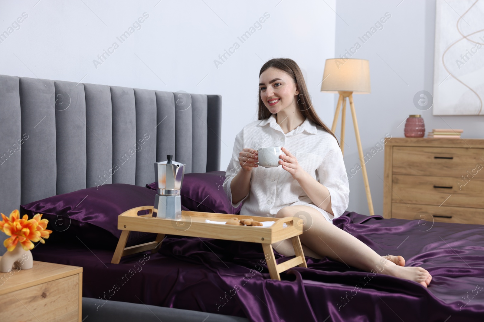 Smiling woman with cup of coffee on bed with purple silk linens indoors Photo of Smiling woman with cup of coffee on bed with purple silk linens indoors