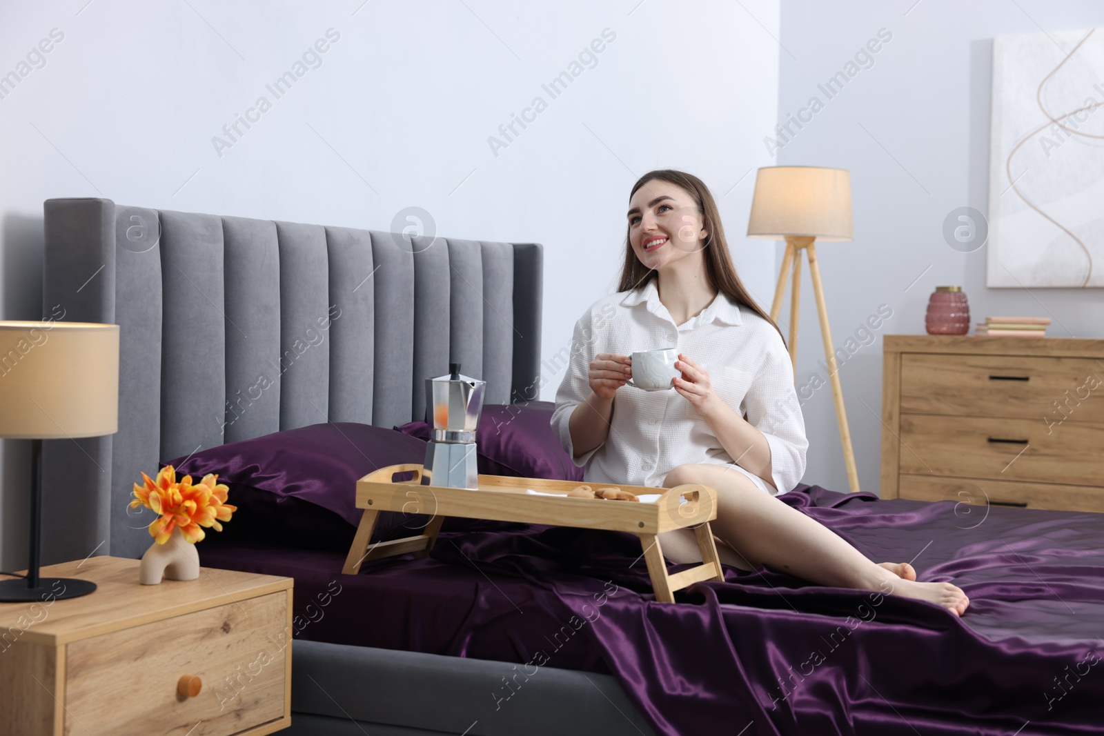 Smiling woman with cup of coffee on bed with purple silk linens indoors Photo of Smiling woman with cup of coffee on bed with purple silk linens indoors