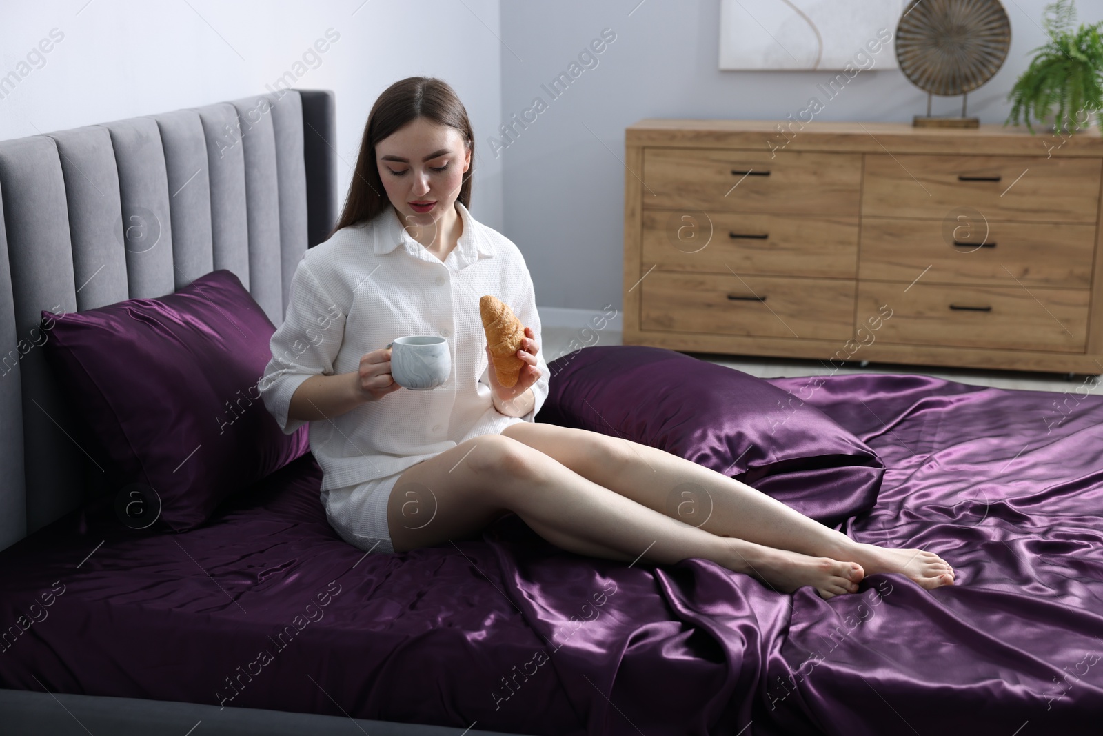 Woman with coffee and croissant on bed with purple silk linens indoors Photo of Woman with coffee and croissant on bed with purple silk linens indoors