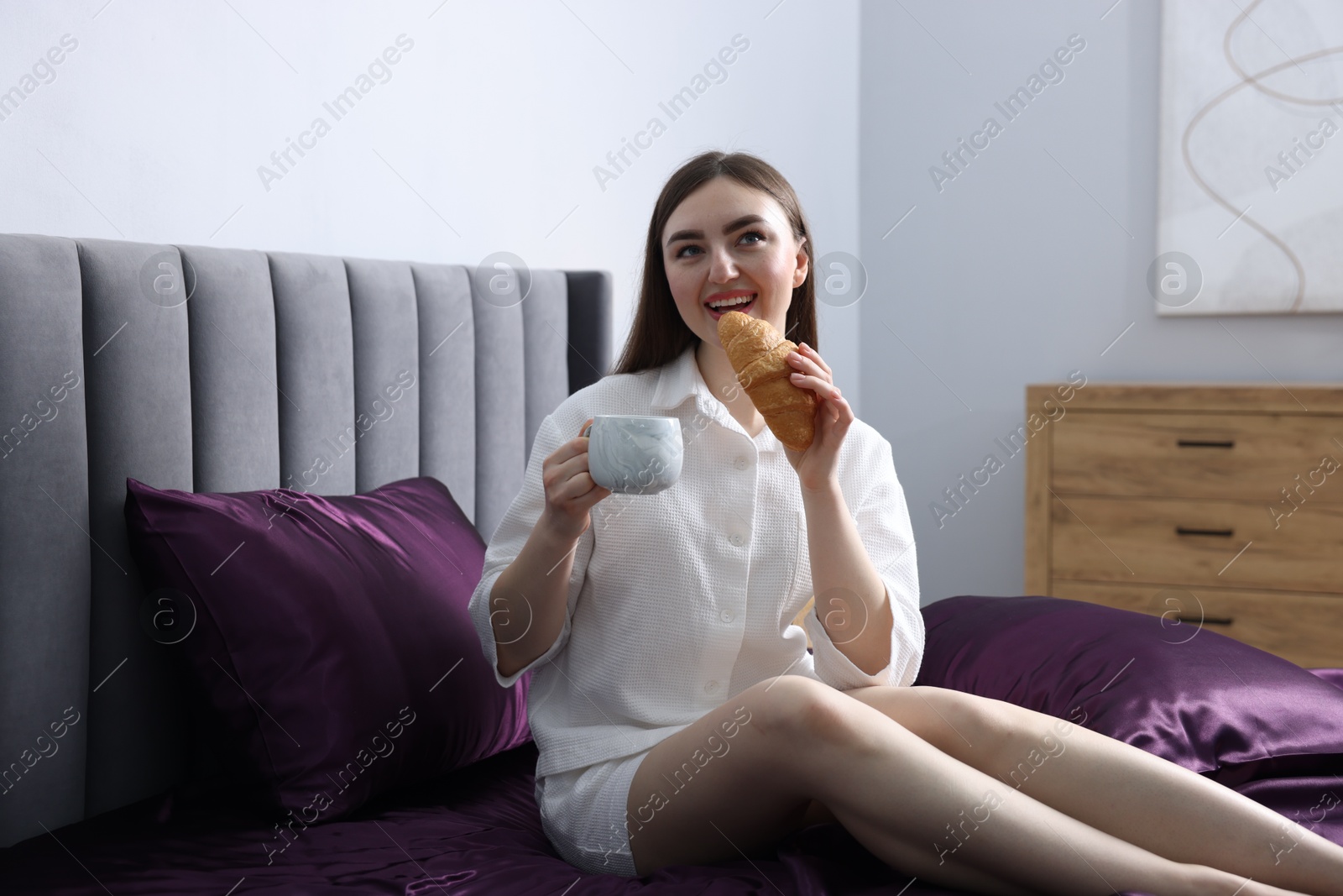 Happy woman with coffee and croissant on bed with purple silk linens indoors Photo of Happy woman with coffee and croissant on bed with purple silk linens indoors