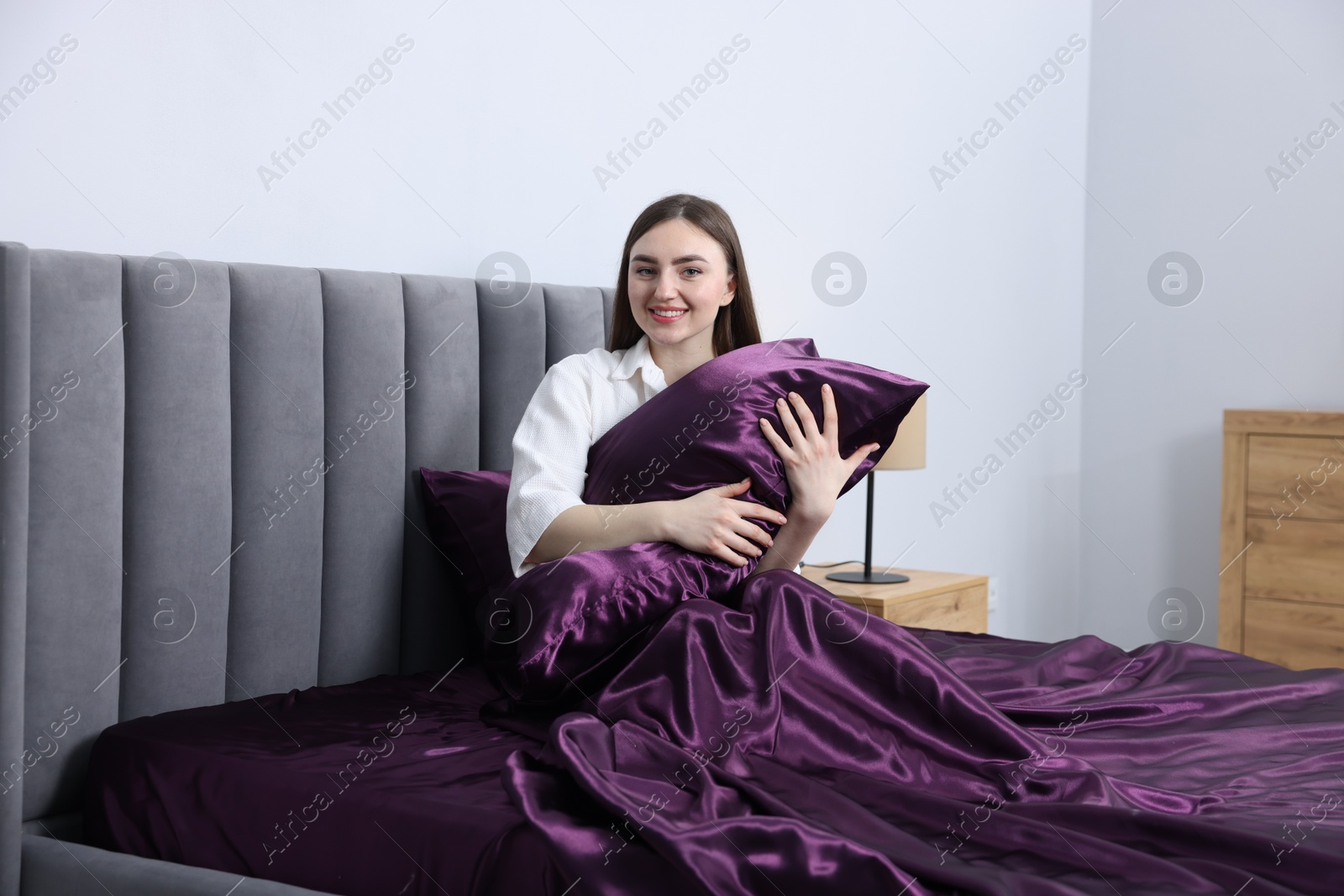 Smiling woman hugging pillow in bed with purple silk linens indoors Photo of Smiling woman hugging pillow in bed with purple silk linens indoors