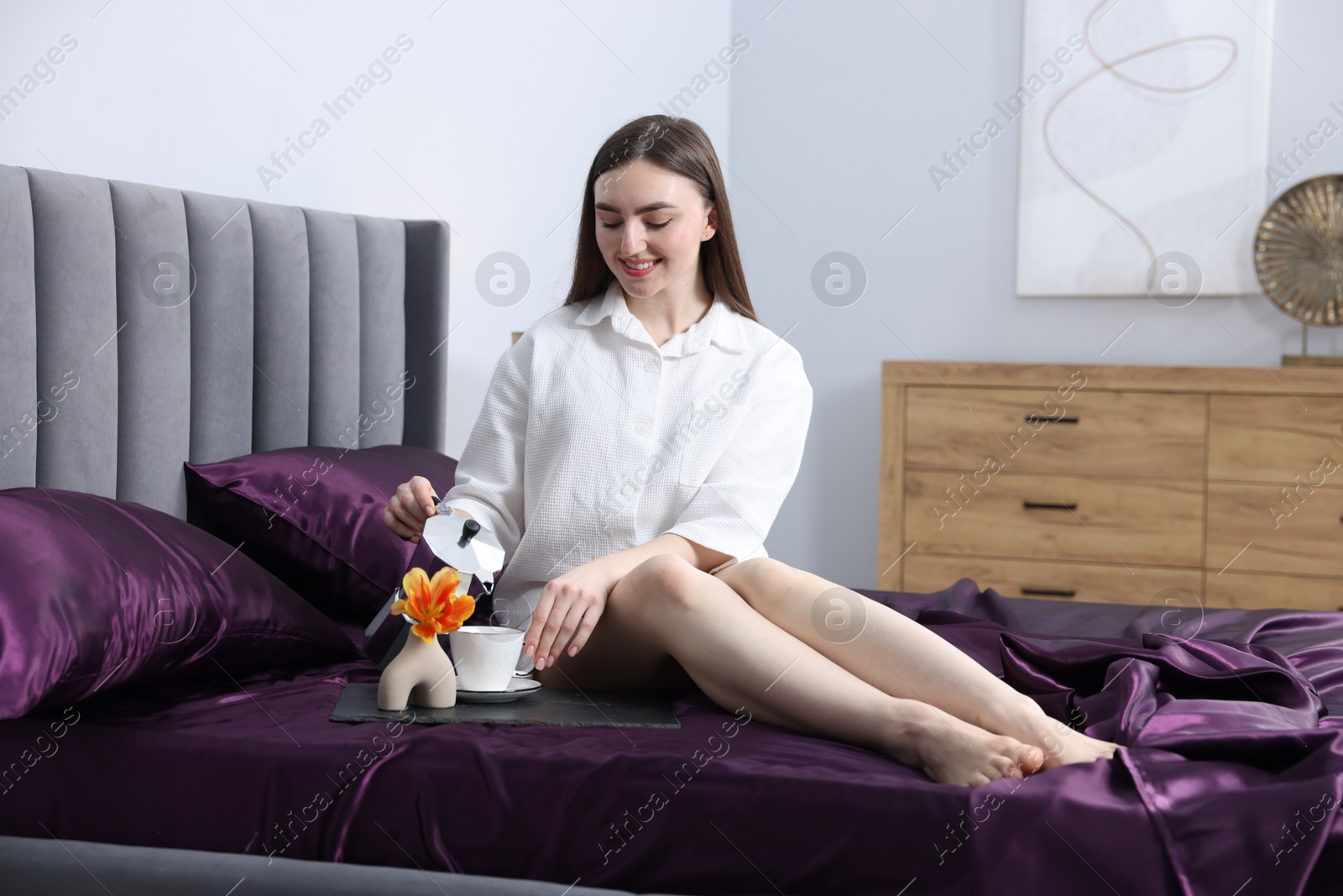 Smiling woman pouring coffee from moka pot into cup on bed with purple silk linens indoors Photo of Smiling woman pouring coffee from moka pot into cup on bed with purple silk linens indoors