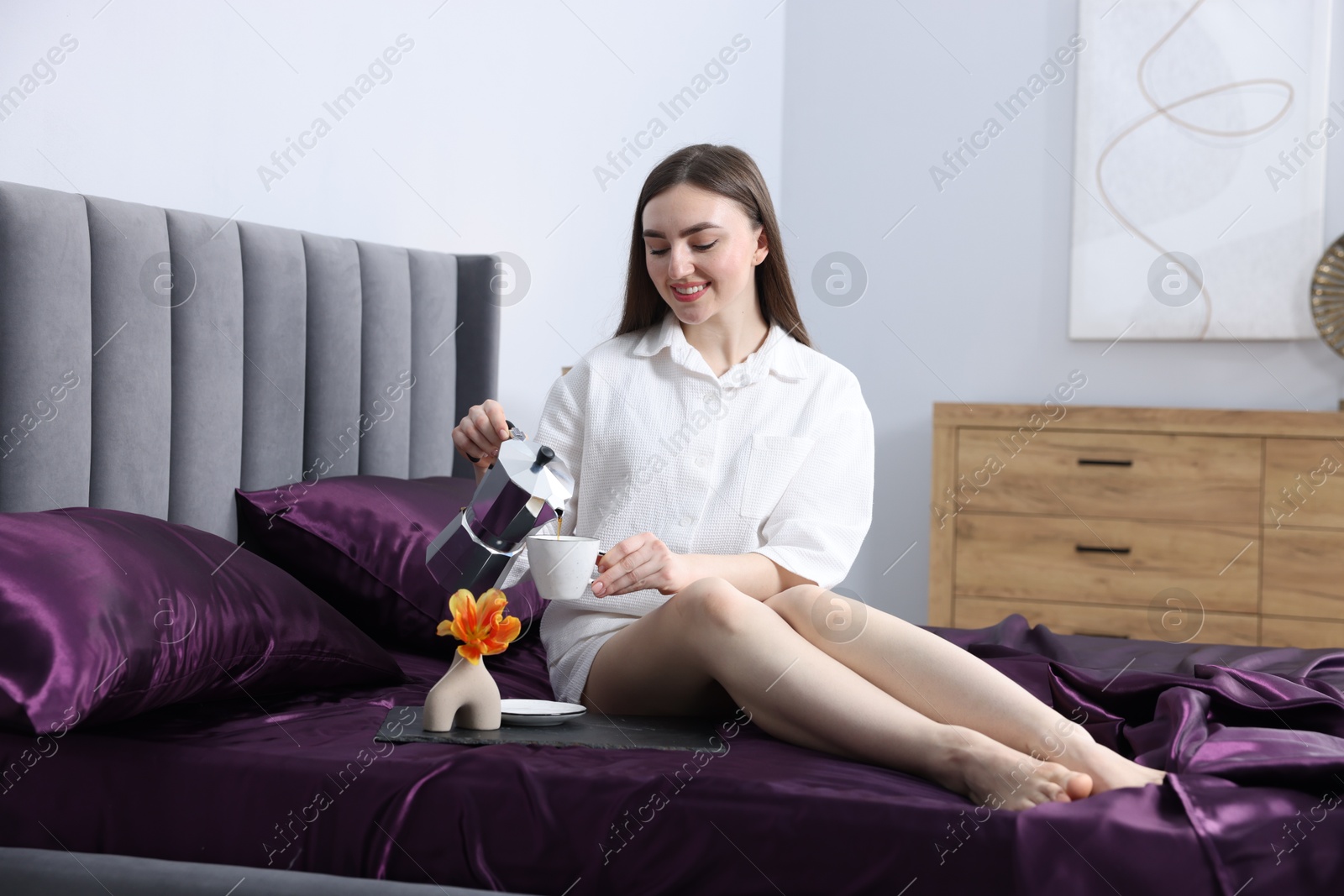Smiling woman pouring coffee from moka pot into cup on bed with purple silk linens indoors Photo of Smiling woman pouring coffee from moka pot into cup on bed with purple silk linens indoors