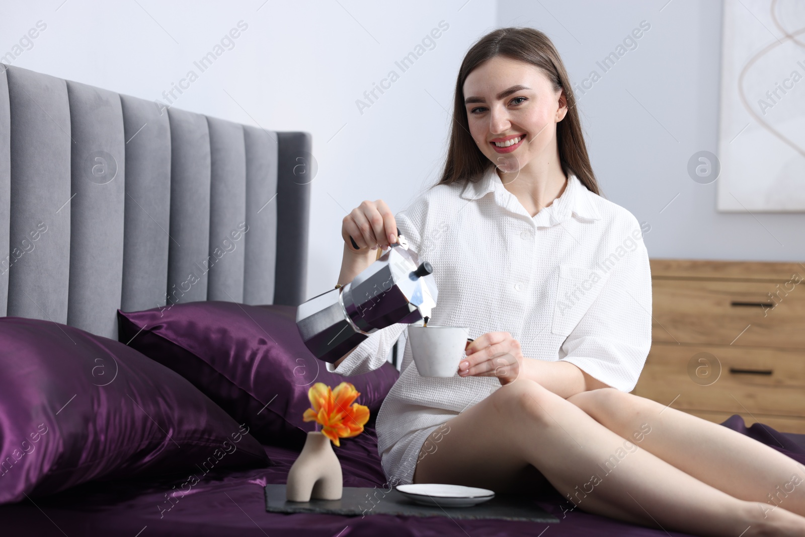 Smiling woman pouring coffee from moka pot into cup on bed with purple silk linens indoors Photo of Smiling woman pouring coffee from moka pot into cup on bed with purple silk linens indoors