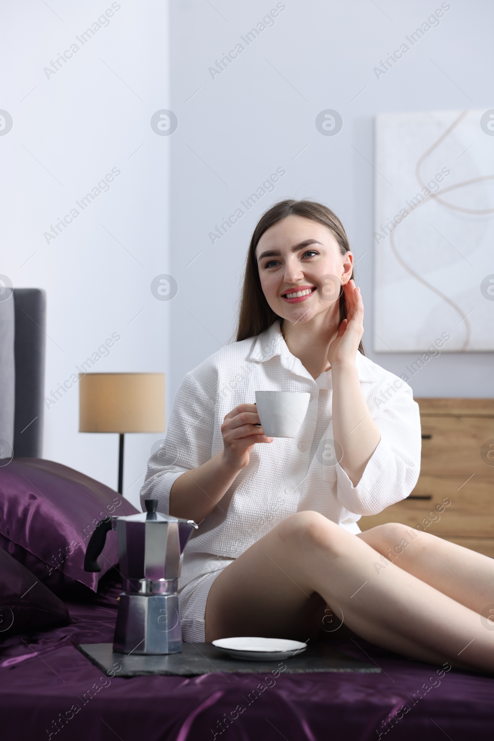 Smiling woman with coffee on bed with purple silk linens indoors Photo of Smiling woman with coffee on bed with purple silk linens indoors