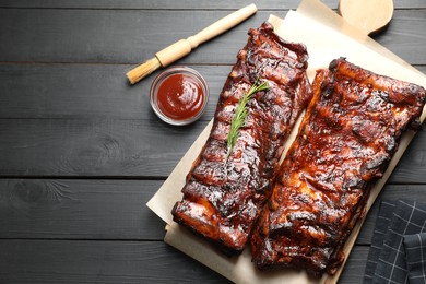 Photo of Tasty roasted pork ribs served with rosemary and sauce on black wooden table, flat lay. Space for text