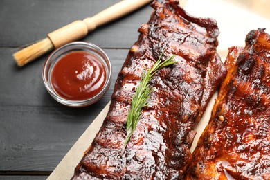 Photo of Tasty roasted pork ribs served with rosemary and sauce on black wooden table, above view