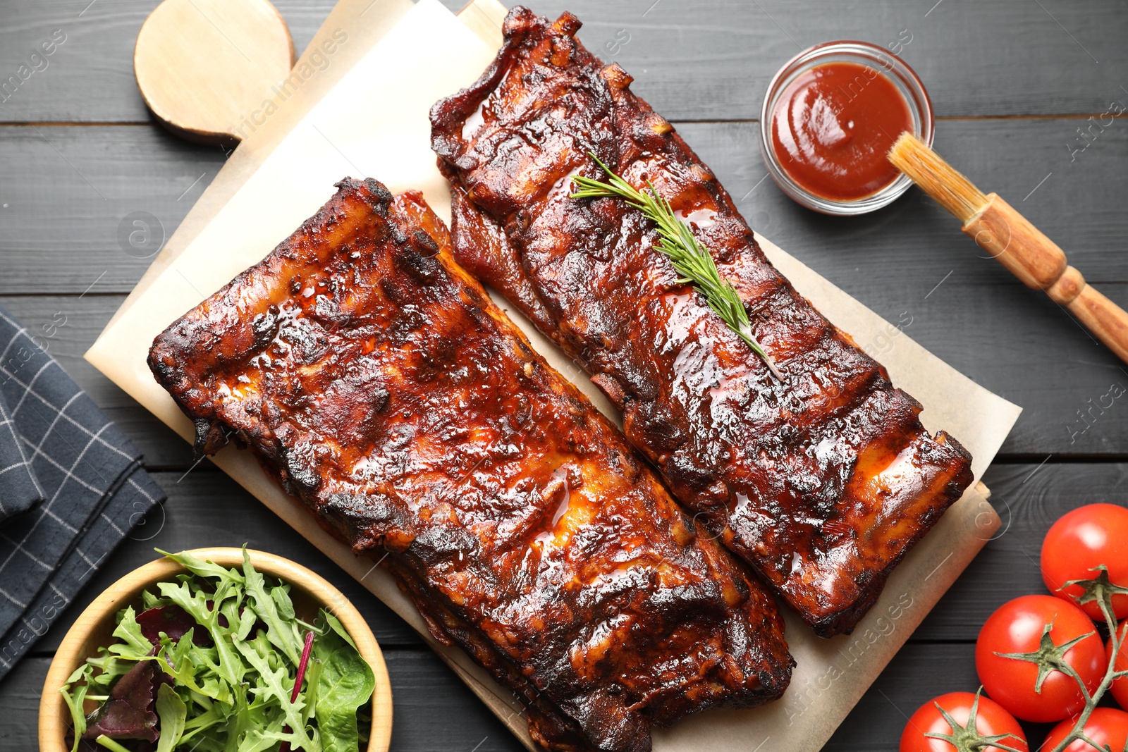 Tasty roasted pork ribs served with rosemary, tomatoes, sauce and salad on black wooden table, flat lay Photo of Tasty roasted pork ribs served with rosemary, tomatoes, sauce and salad on black wooden table, flat lay