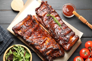 Photo of Tasty roasted pork ribs served with rosemary, tomatoes, sauce and salad on black wooden table, flat lay