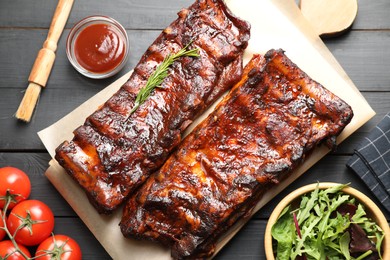Photo of Tasty roasted pork ribs served with rosemary, tomatoes, sauce and salad on black wooden table, flat lay