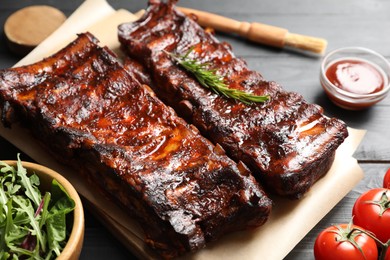 Photo of Tasty roasted pork ribs served with rosemary, tomatoes, sauce and salad on black wooden table, closeup