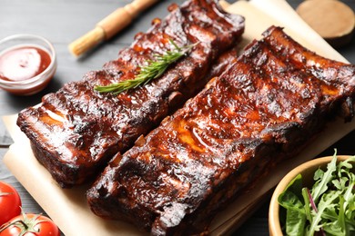 Photo of Tasty roasted pork ribs served with rosemary, tomatoes, sauce and salad on black wooden table, closeup